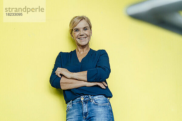 Happy businesswoman with arms crossed leaning on yellow wall