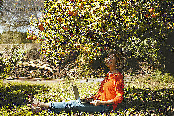 Reife Frau sitzt mit Laptop unter Orangenbaum