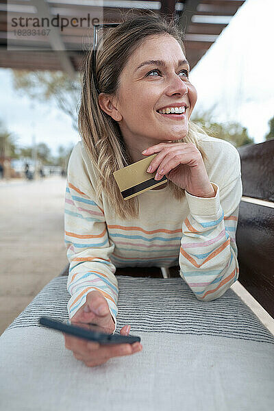 Nachdenkliche junge Frau liegt auf einer Bank und hält Kreditkarte und Smartphone in der Hand