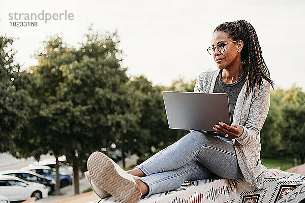 Nachdenkliche reife Frau sitzt bei Sonnenuntergang mit Laptop auf der Terrasse