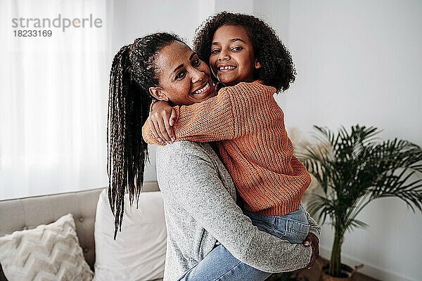 Glückliche Frau trägt und umarmt ihre Tochter im heimischen Wohnzimmer