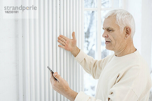 Ein pensionierter älterer Mann überprüft den Heizkörper und hält sein Smartphone zu Hause in der Hand