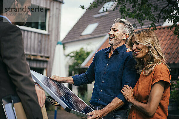Glückliches Paar hält Solarpanel in der Hand und diskutiert mit Immobilienmakler im Hinterhof