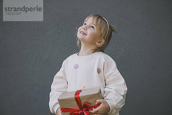 Lächelndes Mädchen schaut nach oben und hält eine Geschenkbox vor der Wand