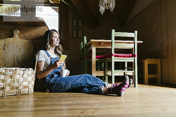 Nachdenkliche Frau mit Kaffeetasse und Smartphone sitzt auf dem Boden