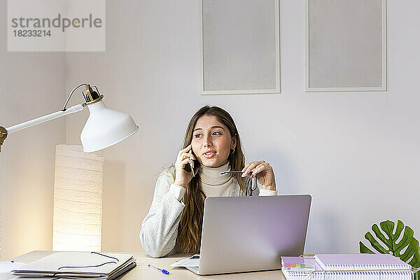 Junge Frau spricht am Schreibtisch mit dem Smartphone
