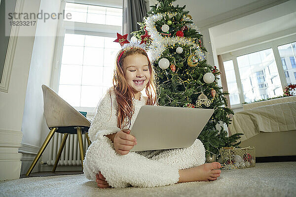 Fröhliches Mädchen mit Laptop sitzt zu Hause vor dem Weihnachtsbaum