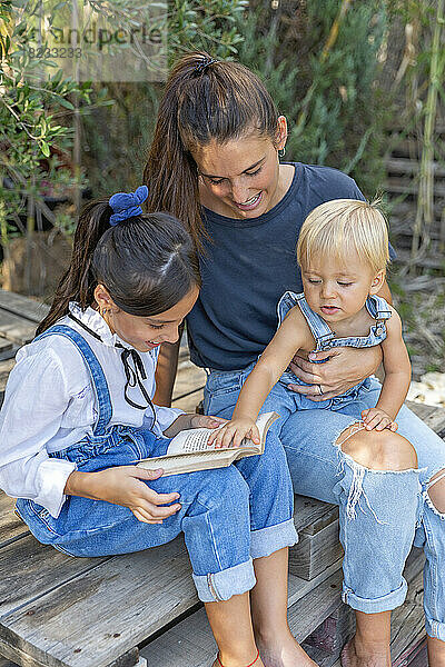 Lächelndes Mädchen liest Buch von Mutter und Bruder  die auf einer Holzpalette sitzen