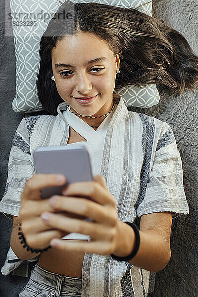 Lächelndes Mädchen benutzt Smartphone  das zu Hause auf dem Teppich liegt