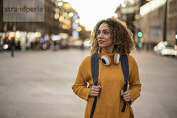 Junge Frau mit Rucksack und kabellosen Kopfhörern geht bei Sonnenuntergang auf Fußweg