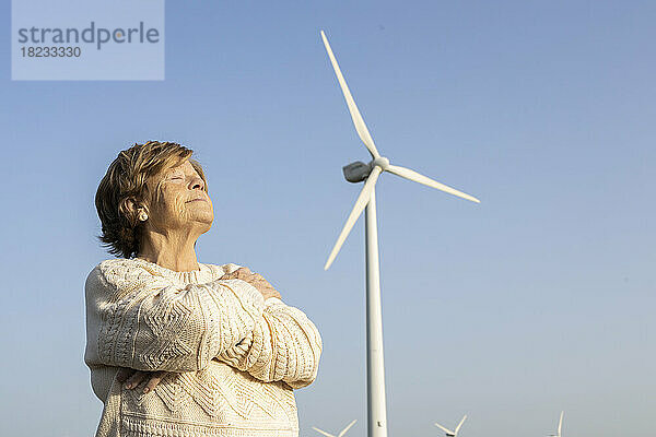 Lächelnde ältere Frau steht mit geschlossenen Augen vor einer Windkraftanlage