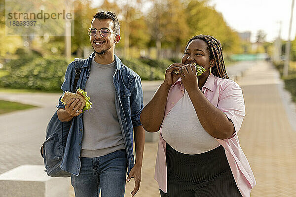 Glückliches junges Paar isst Sandwich und geht auf dem Fußweg