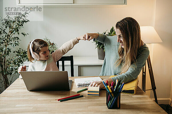 Happy mother giving fist bump to daughter sitting with laptop at table