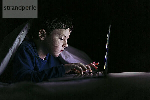 Junge benutzt Laptop und liegt zu Hause unter Decke