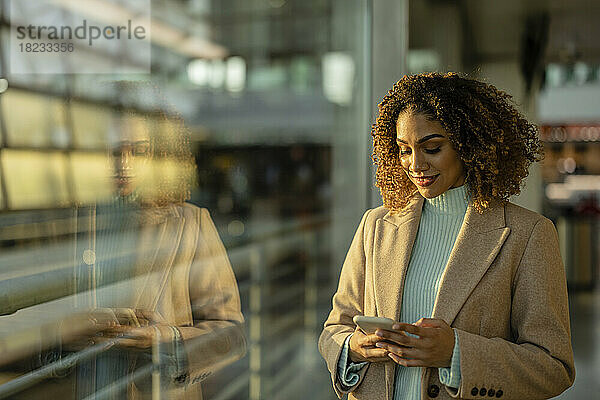 Lächelnde Frau benutzt Smartphone am Fenster mit Reflexion auf Glas am Bahnhof