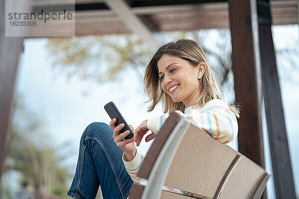Lächelnde schöne junge Frau  die auf einer Bank sitzt und ihr Mobiltelefon benutzt