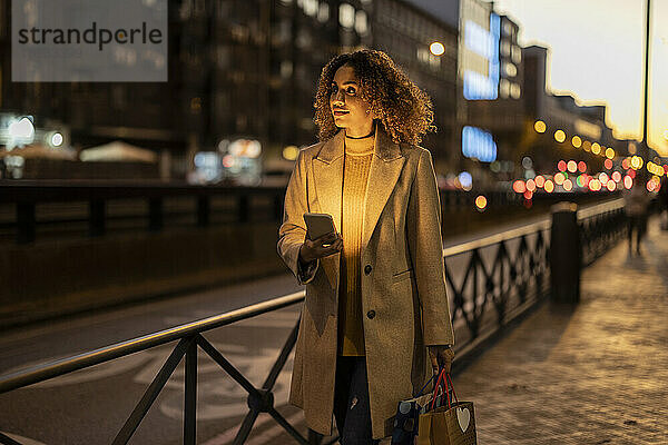 Frau hält Einkaufstüten und Mobiltelefon in der Abenddämmerung auf dem Fußweg
