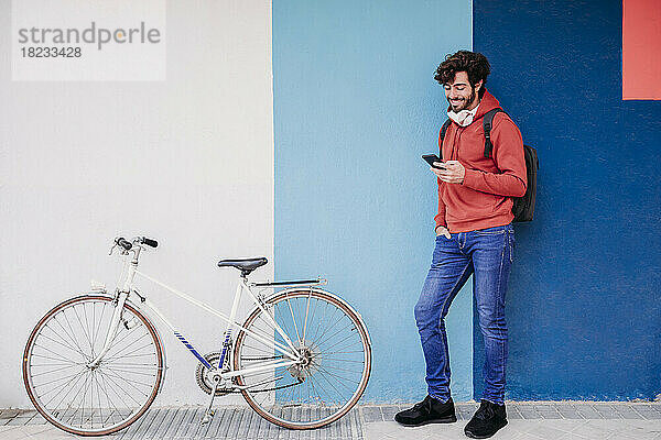 Lächelnder Mann benutzt Smartphone mit dem Fahrrad in der Nähe einer bunten Wand