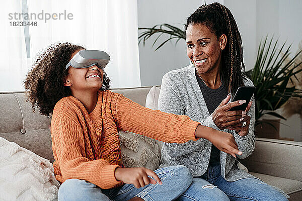 Lächelnde Mutter mit Tochter im Virtual-Reality-Simulator auf dem heimischen Sofa