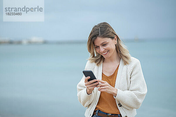Lächelnde schöne junge Frau  die am Strand Textnachrichten auf dem Mobiltelefon sendet