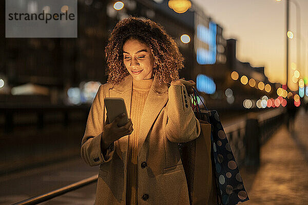 Lächelnde Frau hält in der Abenddämmerung Einkaufstüten mit Smartphone in der Hand