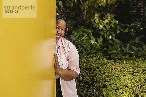 Smiling young woman hiding behind yellow wall