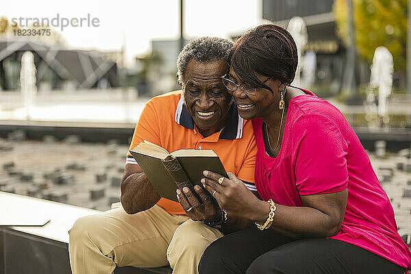 Lächelndes älteres Paar  das an der Wand sitzt und ein Buch teilt