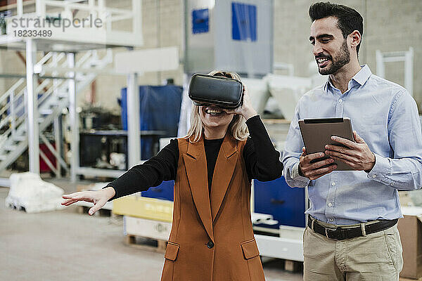 Glücklicher Geschäftsmann hält Tablet-PC in der Hand und steht einem Kollegen mit Virtual-Reality-Simulator zur Seite