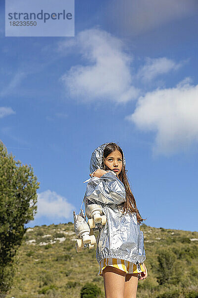 Mädchen in silberner Jacke steht mit Rollschuhen auf einem Berg unter blauem Himmel