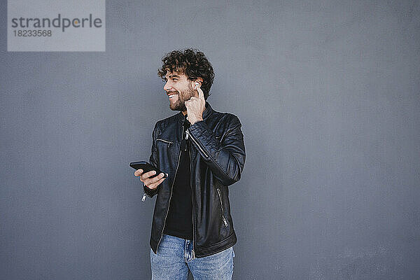 Glücklicher Mann hält sein Mobiltelefon in der Hand und stellt drahtlose In-Ear-Kopfhörer vor einer grauen Wand ein