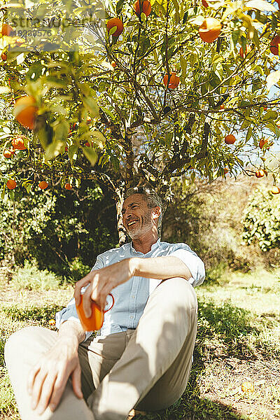 Glücklicher reifer Mann sitzt unter einem Baum mit einer Kaffeetasse im Garten