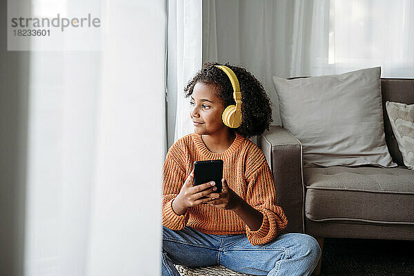 Lächelndes Mädchen mit kabellosen Kopfhörern  das zu Hause mit dem Smartphone sitzt