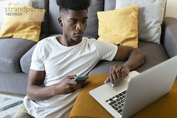 Man with smart phone using laptop sitting near sofa at home