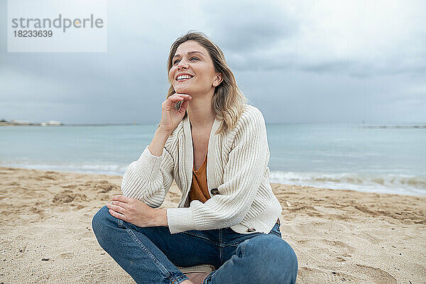 Lächelnde junge schöne Frau sitzt auf Sand und träumt am Strand