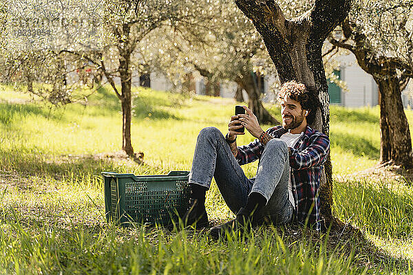 Glücklicher Mann  der Musik über kabellose In-Ear-Kopfhörer hört und ein Smartphone benutzt  das an einen Baum lehnt