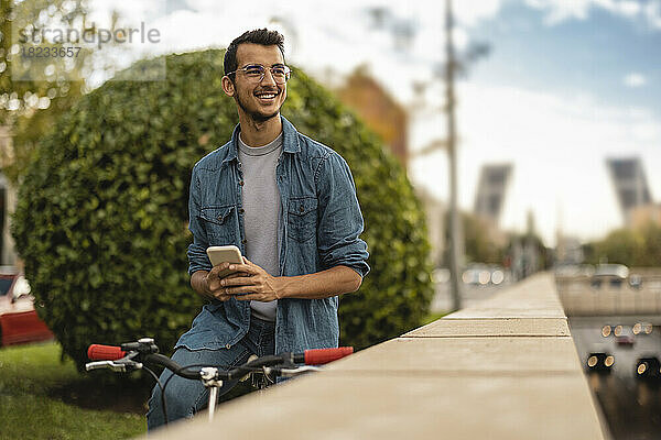 Glücklicher junger Mann mit Smartphone  der auf dem Fahrrad sitzt