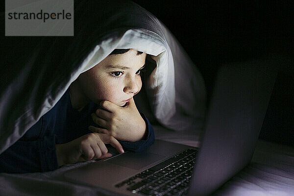 Nachdenklicher Junge schaut auf den Laptop  der unter der Decke auf dem Bett liegt