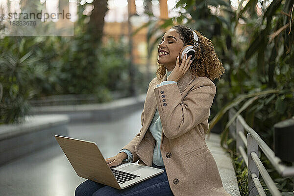 Glückliche Geschäftsfrau  die Musik über drahtlose Kopfhörer hört und mit einem Laptop auf der Bank sitzt