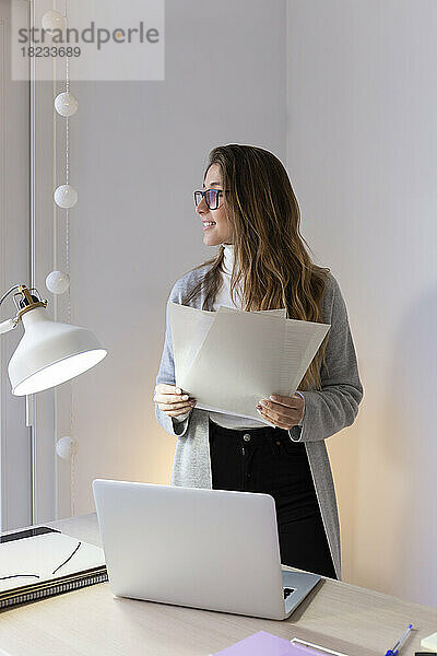 Smiling young woman standing with documents at desk