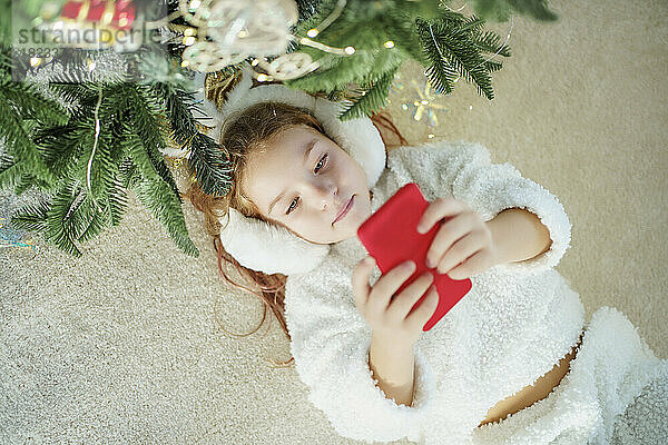 Mädchen liegt auf Teppich neben Weihnachtsbaum und benutzt Smartphone zu Hause