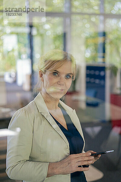 Reife Geschäftsfrau hält Mobiltelefon im Büro  gesehen durch Glas