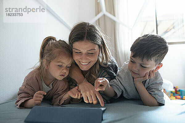 Lächelnde junge Mutter mit Kindern  die zu Hause im Schlafzimmer einen Tablet-PC nutzt