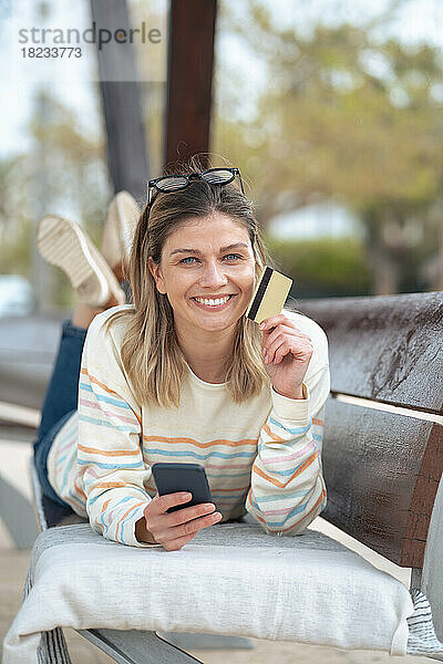 Lächelnde junge Frau liegt auf einer Bank und hält Kreditkarte und Smartphone in der Hand