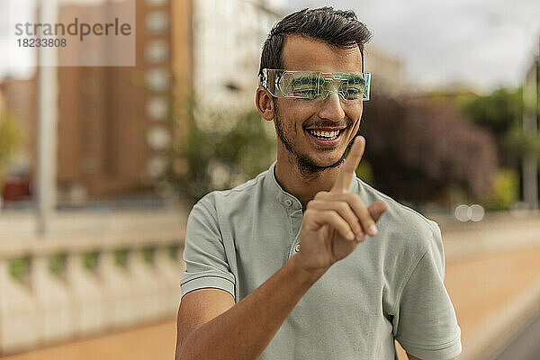 Glücklicher junger Mann gestikuliert mit einer Smart-Brille am Fußweg