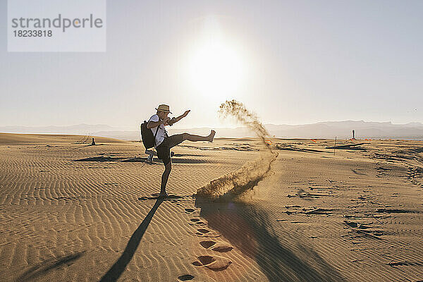 Verspielter Mann kickt Sand am Strand bei Sonnenuntergang
