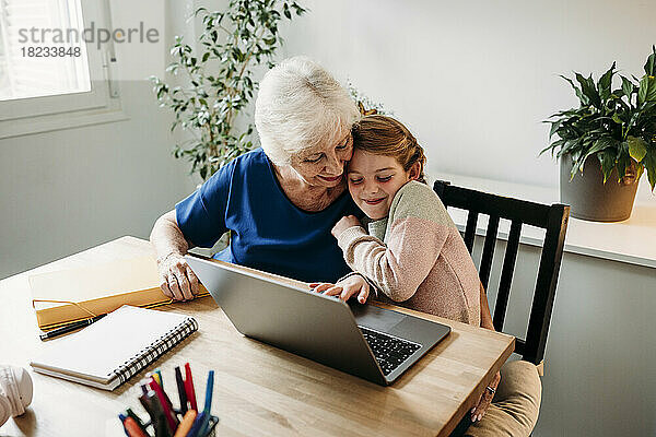 Lächelndes Mädchen mit Laptop auf dem Tisch  das Großmutter zu Hause umarmt