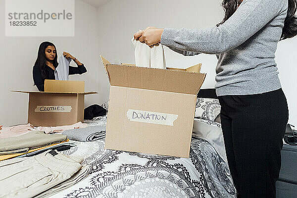 Junge Frauen packen zu Hause auf dem Bett eine Kiste mit Kleidung für eine Spende