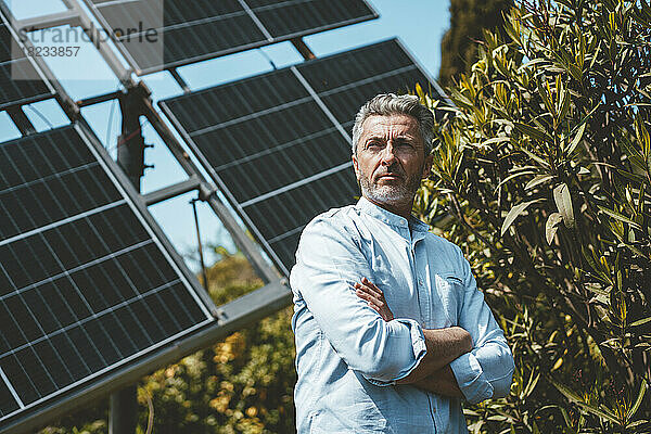 Nachdenklicher Mann mit verschränkten Armen vor Sonnenkollektoren an einem sonnigen Tag