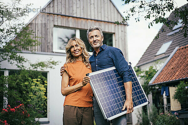 Lächelndes älteres Paar  das mit Solarpanel vor dem Haus steht