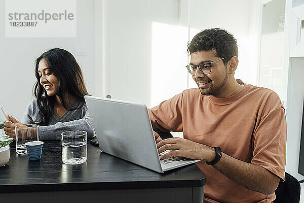 Lächelnder Mann lernt am Laptop und sitzt neben einer Frau am Tisch im Wohnzimmer